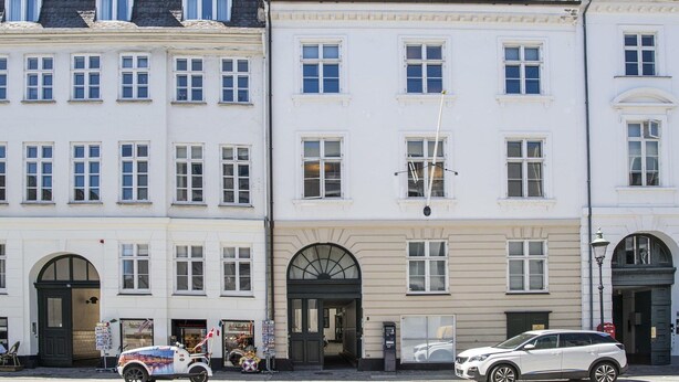 Eine Reihe weißer Gebäude mit zahlreichen Fenstern, ein zentraler Eingang mit Bogen und eine Straße mit geparkten Autos.