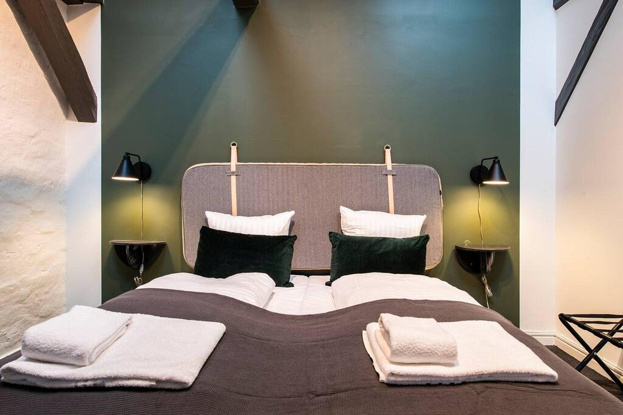 A neatly made bed with pillows and folded towels, two bedside lamps, and a headboard with a light fixture.