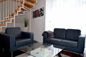 Una sala de estar moderna con una mesa de centro de vidrio, dos sofás, una escalera con escalones de madera, una planta en maceta y una obra de arte abstracto en la pared.