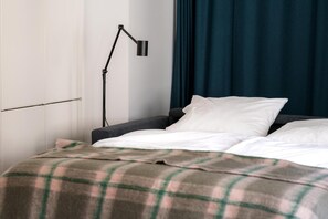 Una cama bien hecha con una manta escocesa, una lámpara de noche y una cortina oscura en una habitación con paredes blancas.
