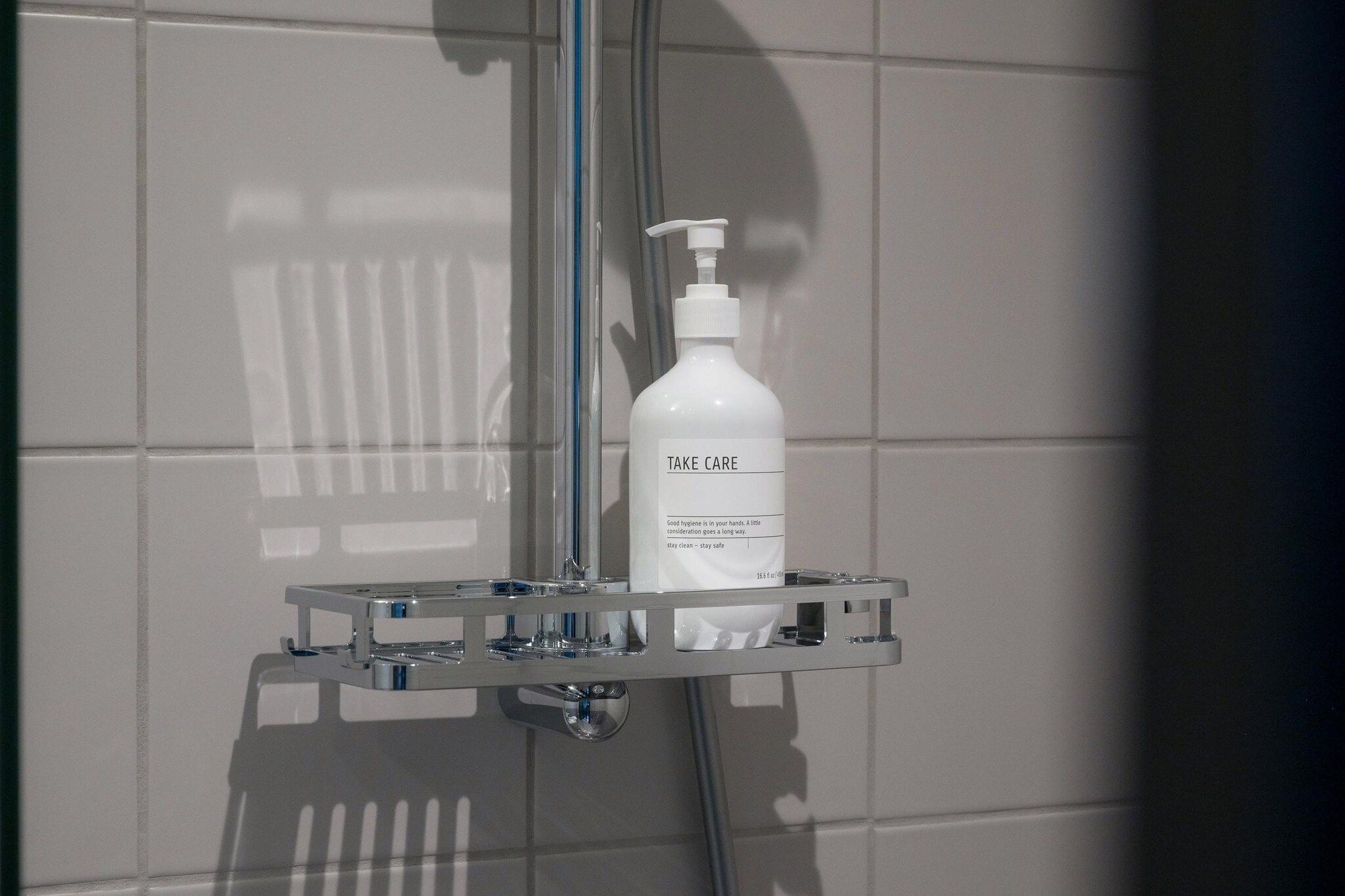 Une salle de bain avec un mur recouvert de carreaux blancs, un receveur de douche en chrome et un flacon de savon liquide sur lequel est inscrit « PRENEZ SOIN DE VOUS ».
