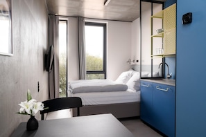 Quarto compacto com cama, escrivaninha e cadeira. Há uma janela com cortinas, uma TV na parede e uma prateleira com objetos.