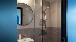 Et moderne badeværelse med en brusekabine i glas, et rundt spejl og en hvid vask med toiletartikler.