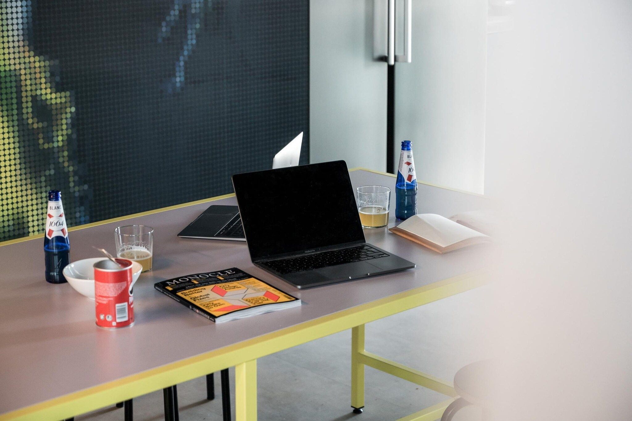 A laptop on a table with a book, a glass of beer, a bottle of water, and a can of soda.
