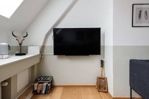 A room with a flat-screen TV mounted on the wall, a wooden floor, a desk with a coffee machine, a skull decoration, and a framed picture of feathers. - Family Retreat Near Kongens Nytorv (København)