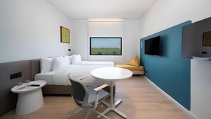 A modern hotel room with a bed, a desk, a chair, a small table, a TV, and a window with a view of a field. - Go Hotel Ban Chang (Ban Chang)