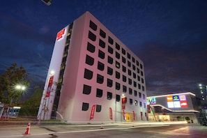 A modern hotel building with a distinctive pink facade, multiple windows, and a prominent red logo on the side. - Go Hotel Ban Chang (Ban Chang)