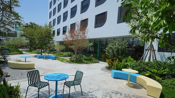 An outdoor seating area with round tables and chairs, surrounded by greenery and a building with large windows. - Go Hotel Ban Chang (Ban Chang)