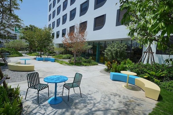 An outdoor seating area with round tables and chairs, surrounded by greenery and a building with large windows. - Go Hotel Ban Chang (Ban Chang)