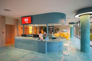 A hotel reception area with a curved blue counter, a digital display, and a seating area with yellow chairs. - Go Hotel Ban Chang (Ban Chang)