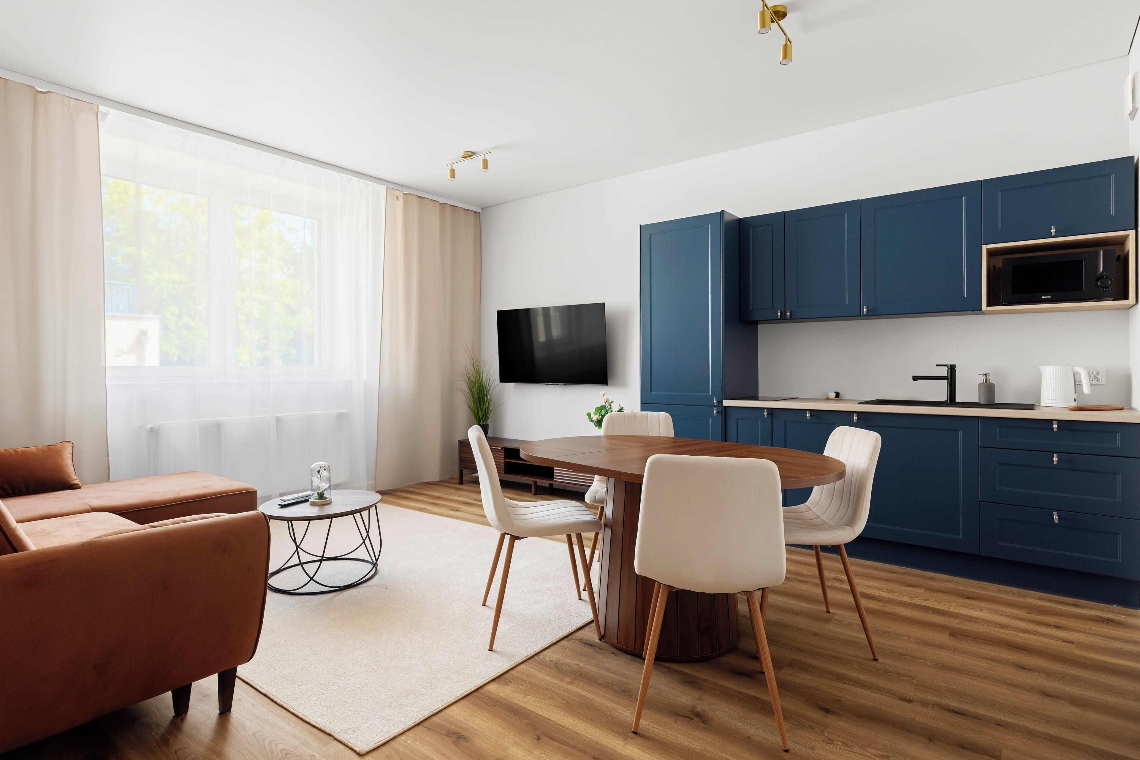 Una sala de estar moderna con un sofá, una mesa de comedor y sillas. Hay una zona de cocina con armarios azules y un microondas.