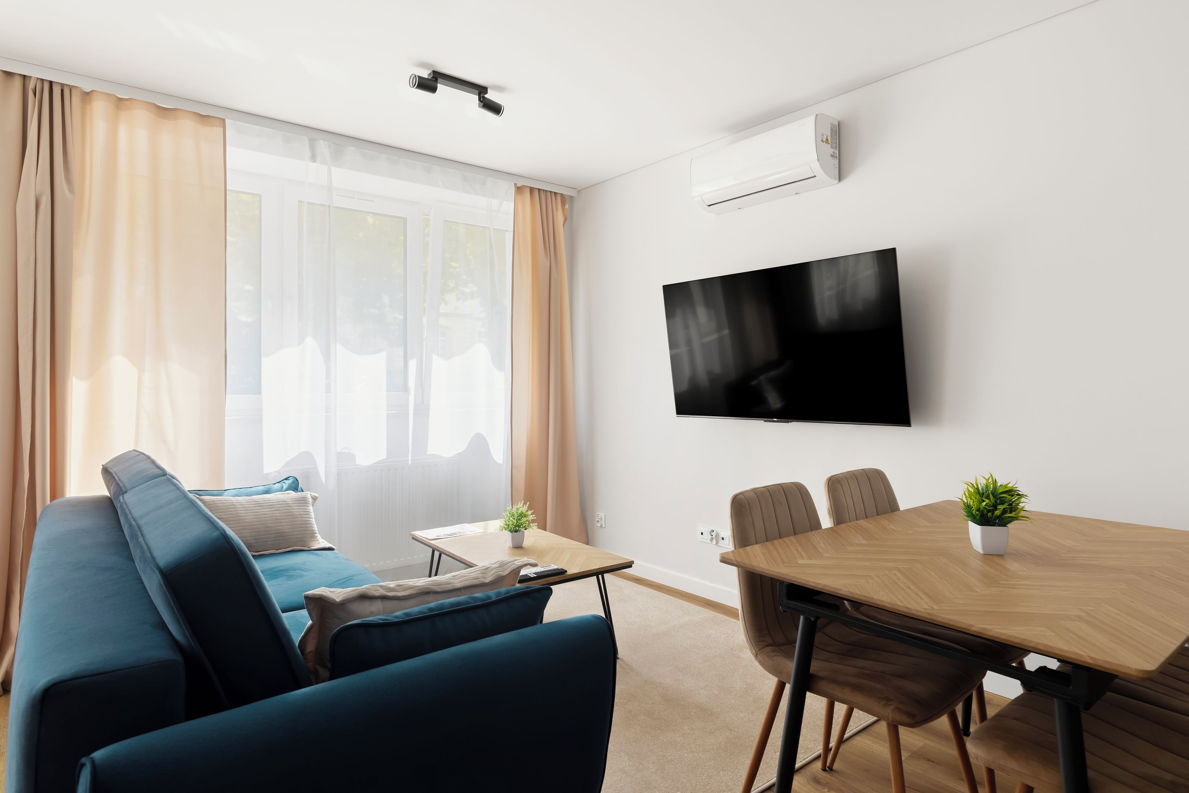 Una sala de estar moderna con sofá, una mesa de centro, una mesa de comedor con sillas, un televisor de pantalla plana montado en la pared y una ventana con cortinas.