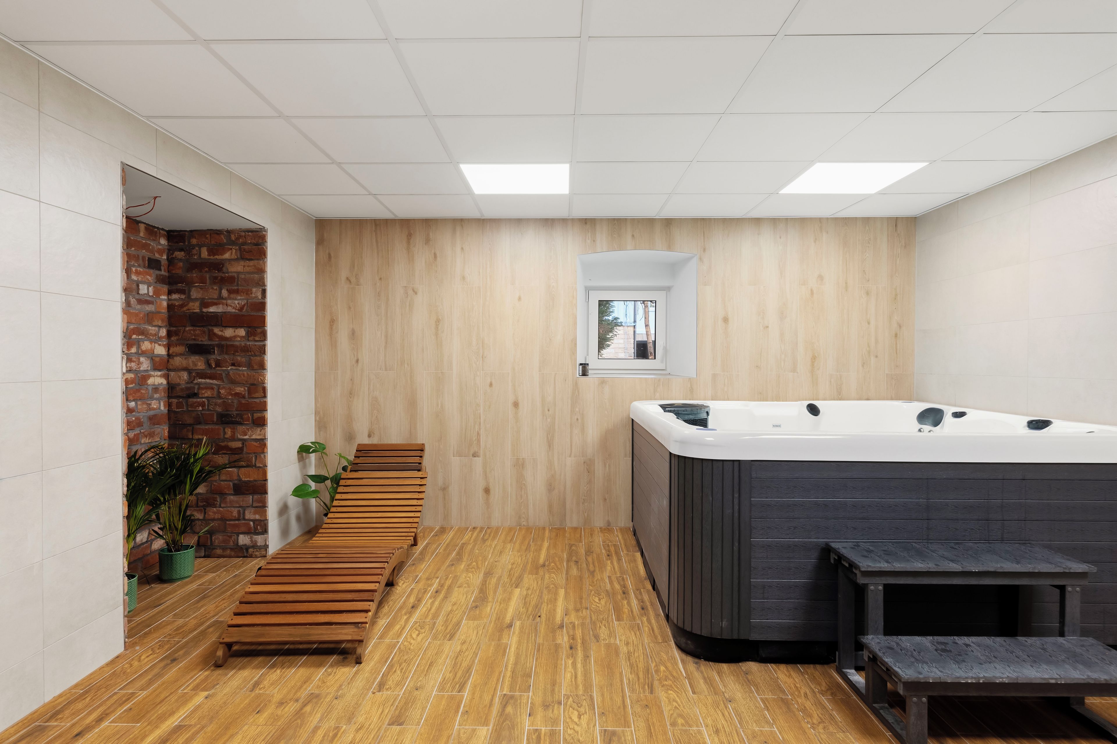 A modern bathroom with a wooden floor, a brick accent wall, a wooden bench, and a hot tub.