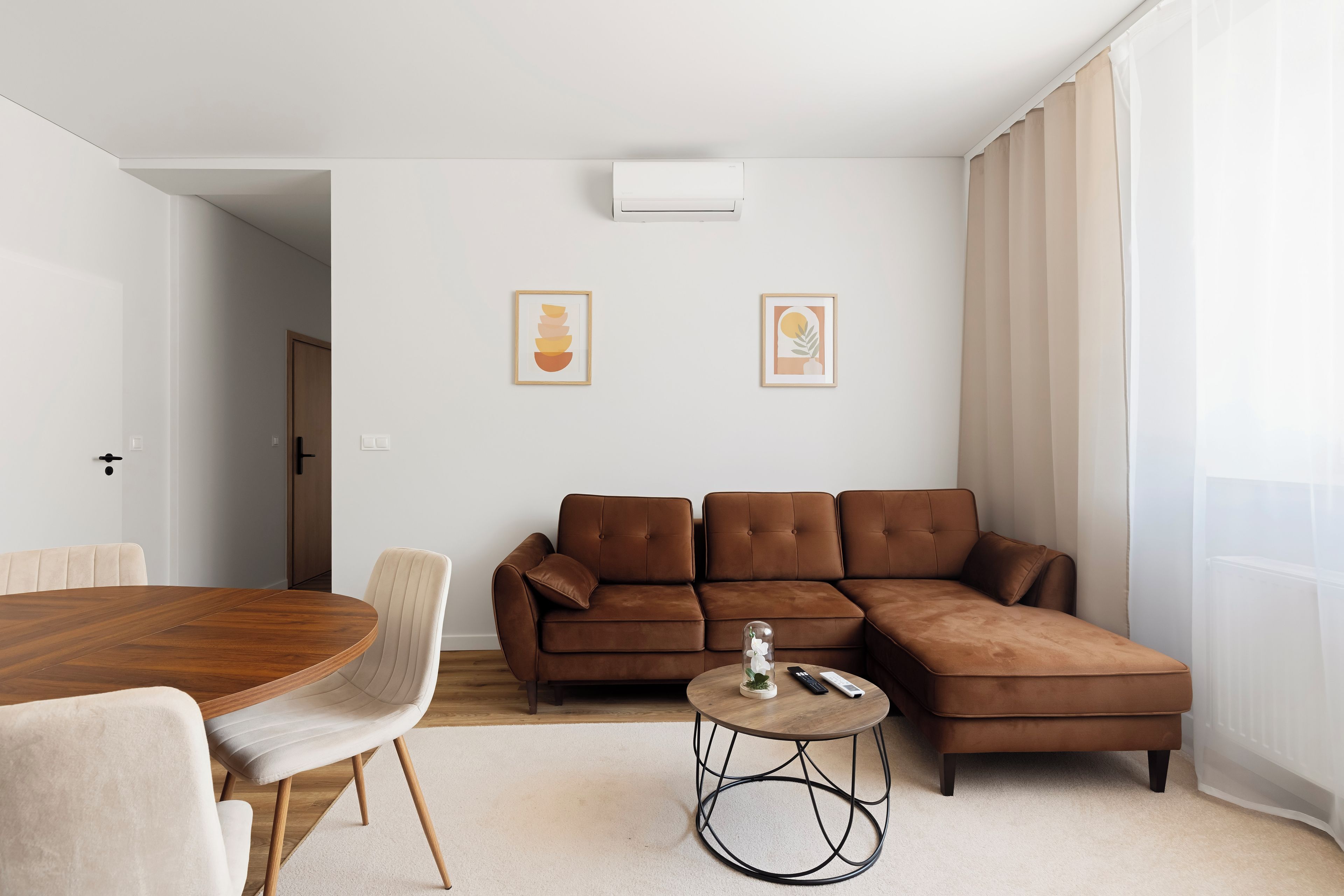 A modern living room with a brown sectional sofa, a round coffee table, and a dining table with chairs. There are two framed artworks on the wall.