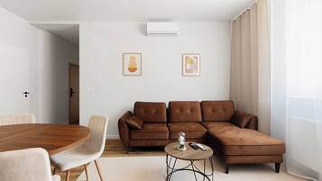 A modern living room with a brown sectional sofa, a round coffee table, and a dining table with chairs. There are two framed artworks on the wall.