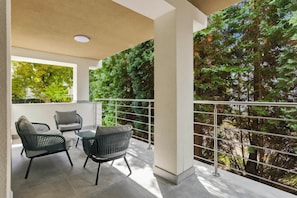 A balcony with a view of trees, a glass railing, and two wicker chairs with cushions.