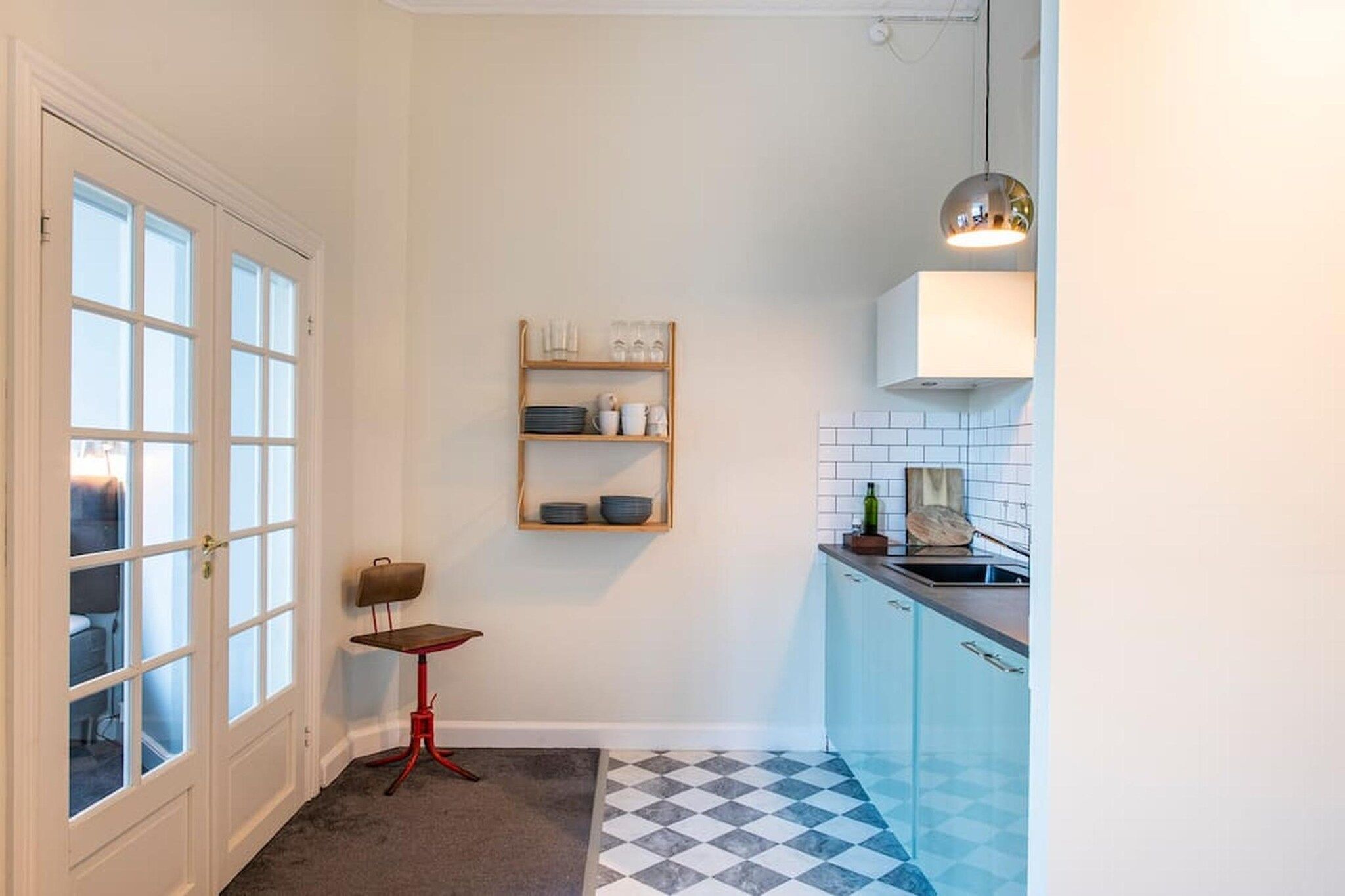 A compact kitchen with a checkered floor, a small table, and a shelf with kitchenware.