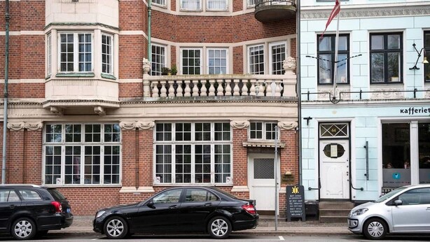Une vue de rue avec une voiture noire garée devant un immeuble dont la porte est blanche et qui porte une enseigne.