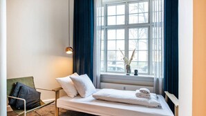 Une chambre d’hôtel avec un lit simple, une chaise, une fenêtre avec des rideaux et un luminaire suspendu.