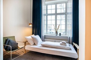 Quarto de hotel com uma cama de solteiro, uma cadeira, uma janela com cortinas e um lustre.