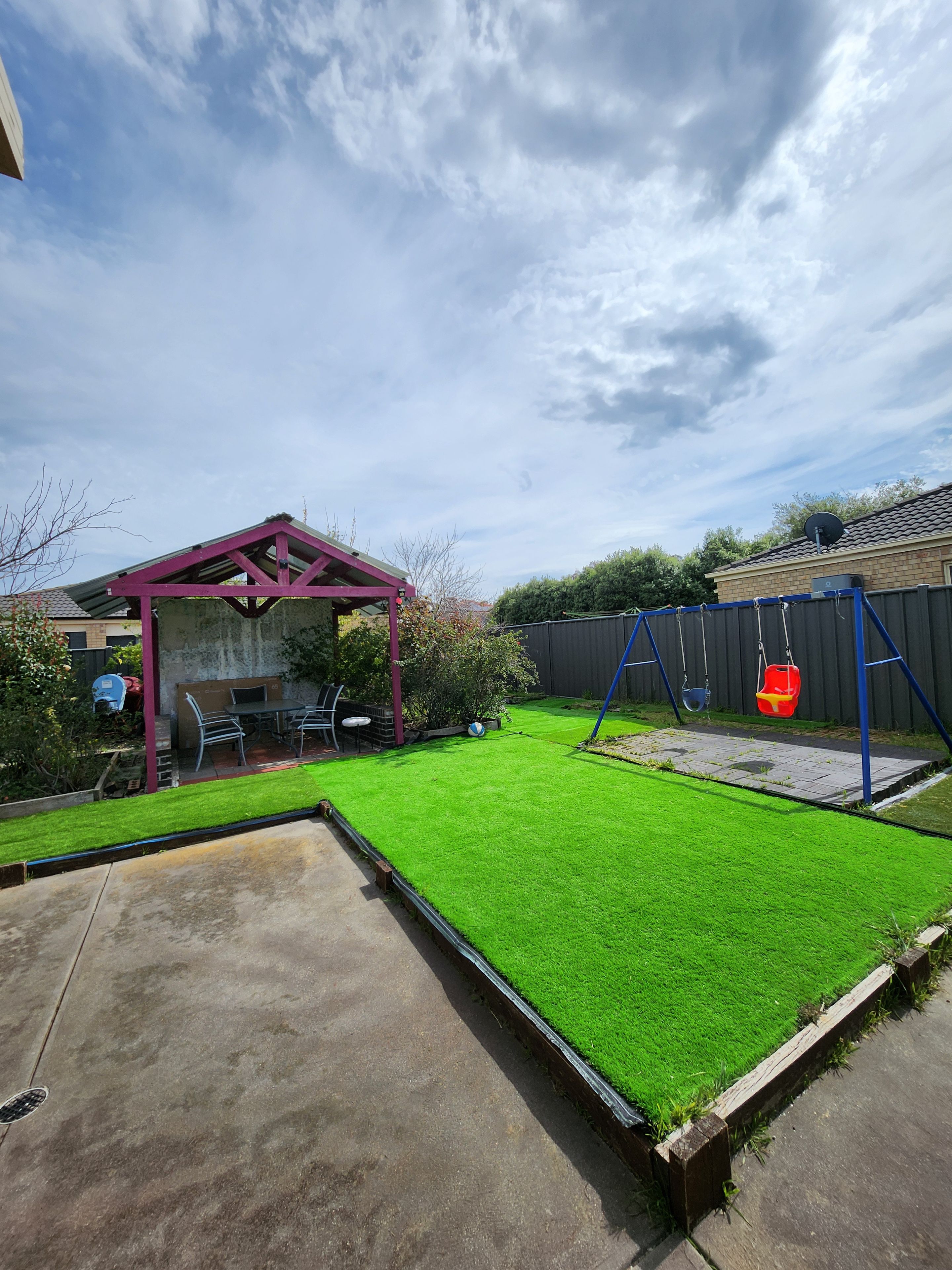 Een achtertuin met kunstgras, een schommelset, een overdekte patio en een houten omheining.
