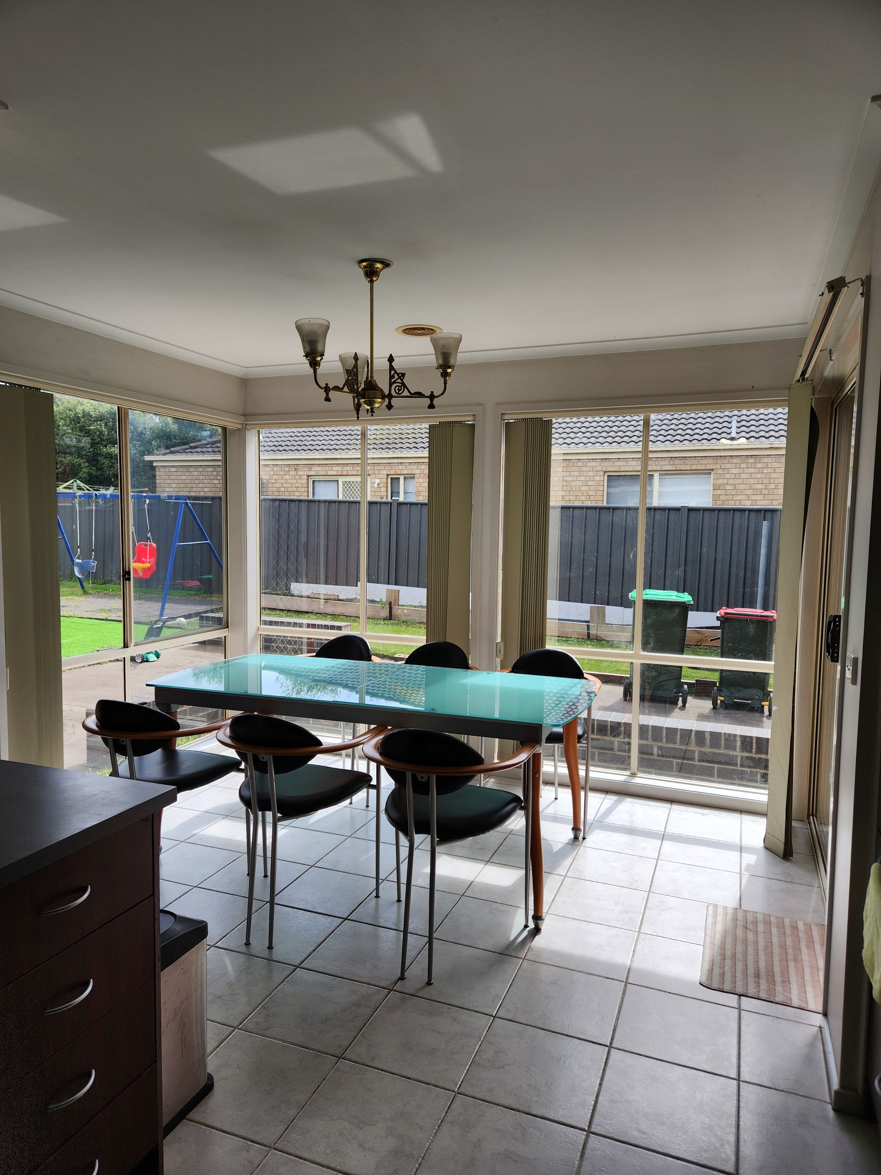 A dining area with a glass table and chairs, a chandelier, and a view of the backyard through large windows.