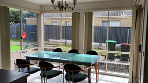 Un comedor con mesa y sillas de vidrio, una araña y vista al patio trasero a través de amplios ventanales.