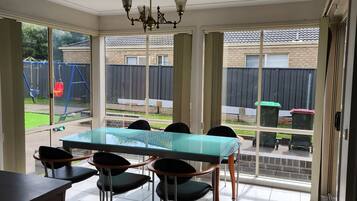 A dining area with a glass table and chairs, a chandelier, and a view of the backyard through large windows.