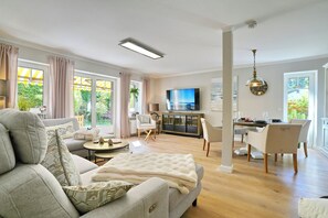 Ein geräumiges Wohnzimmer mit einer Couch, einem Essbereich mit Stühlen, einem Fernseher und Blick auf Grünfläche.
