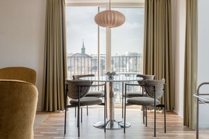 A modern dining area with a round table and four chairs, a hanging light fixture, and a view of a cityscape through large windows. - Coastal flat in Amager with balcony (København)
