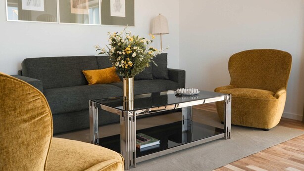 Una sala de estar con un sofá gris, una mesa de centro y dos sillones. Hay cuadros enmarcados en la pared y un florero con flores sobre la mesa de centro.