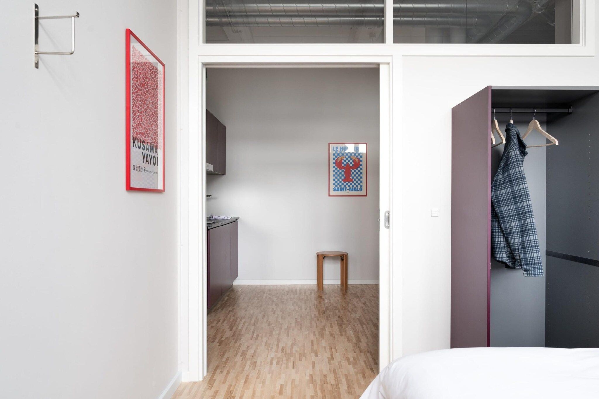 A bedroom with a bed, a wooden floor, a wardrobe, and a door leading to a kitchen.