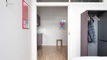 A bedroom with a bed, a wooden floor, a wardrobe, and a door leading to a kitchen.