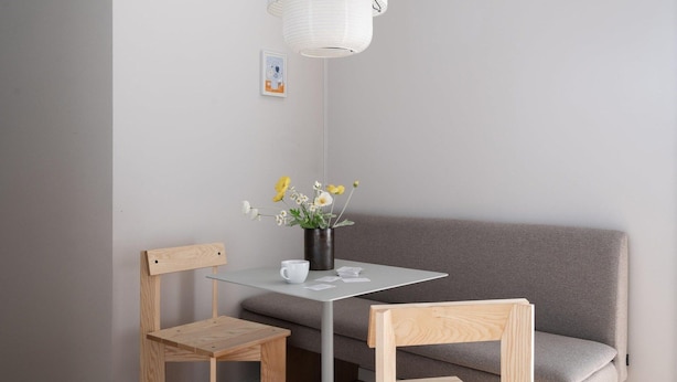 Ein kleiner, moderner Raum mit einem grauen Sofa, zwei Holzstühlen, einem Glastisch und einer weißen Pendelleuchte.