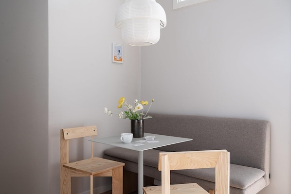 Habitació petita i moderna amb un sofà gris, dues cadires de fusta, una taula de vidre i una làmpada penjant blanca.