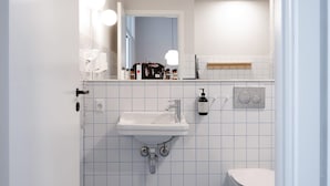 Ein Badezimmer mit einem weißen Waschbecken, einem Spiegel und einer Toilette. Auf dem Waschbeckenbrett liegen Pflegeprodukte.