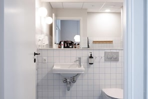 Un bany amb un lavabo blanc, un mirall i un vàter. Hi ha productes de tocador damunt del taulell del lavabo.