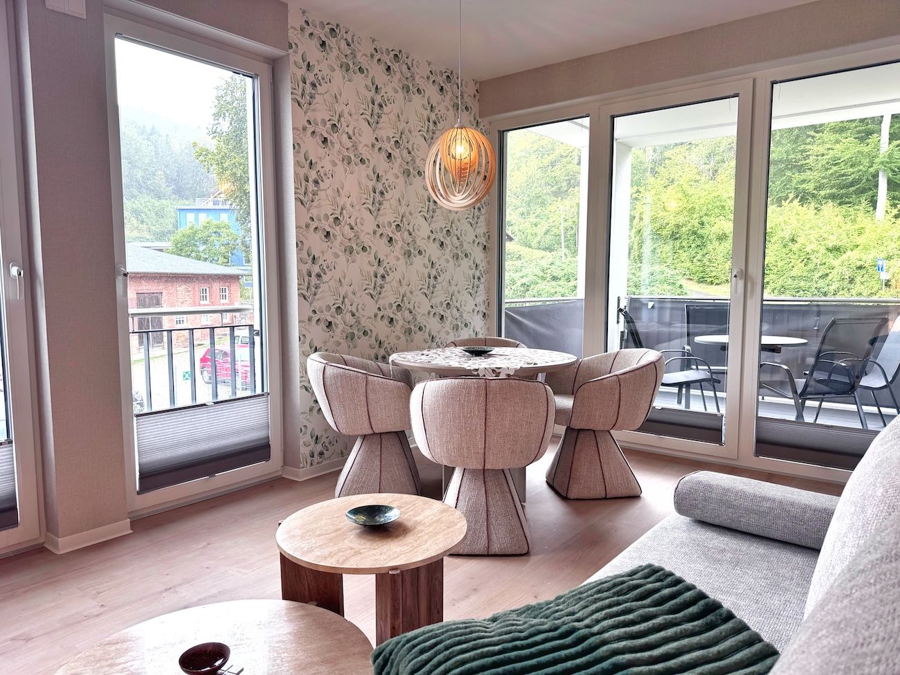 A modern living room with a round table and chairs, large windows with a view of greenery, and a cozy seating area.
