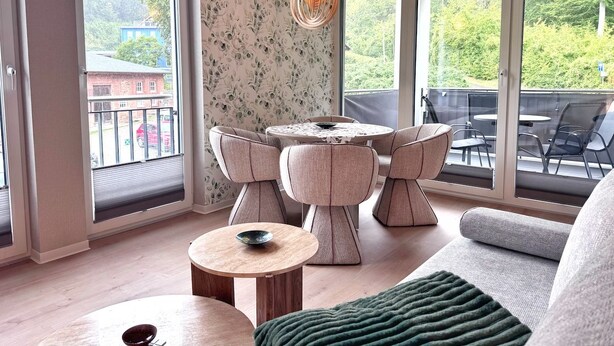 Uma sala de estar moderna com uma mesa redonda e cadeiras, janelas amplas com vista para o verde, e um espaço de estar acolhedor.