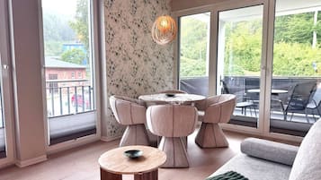 A modern living room with a round table and chairs, large windows with a view of greenery, and a cozy seating area.