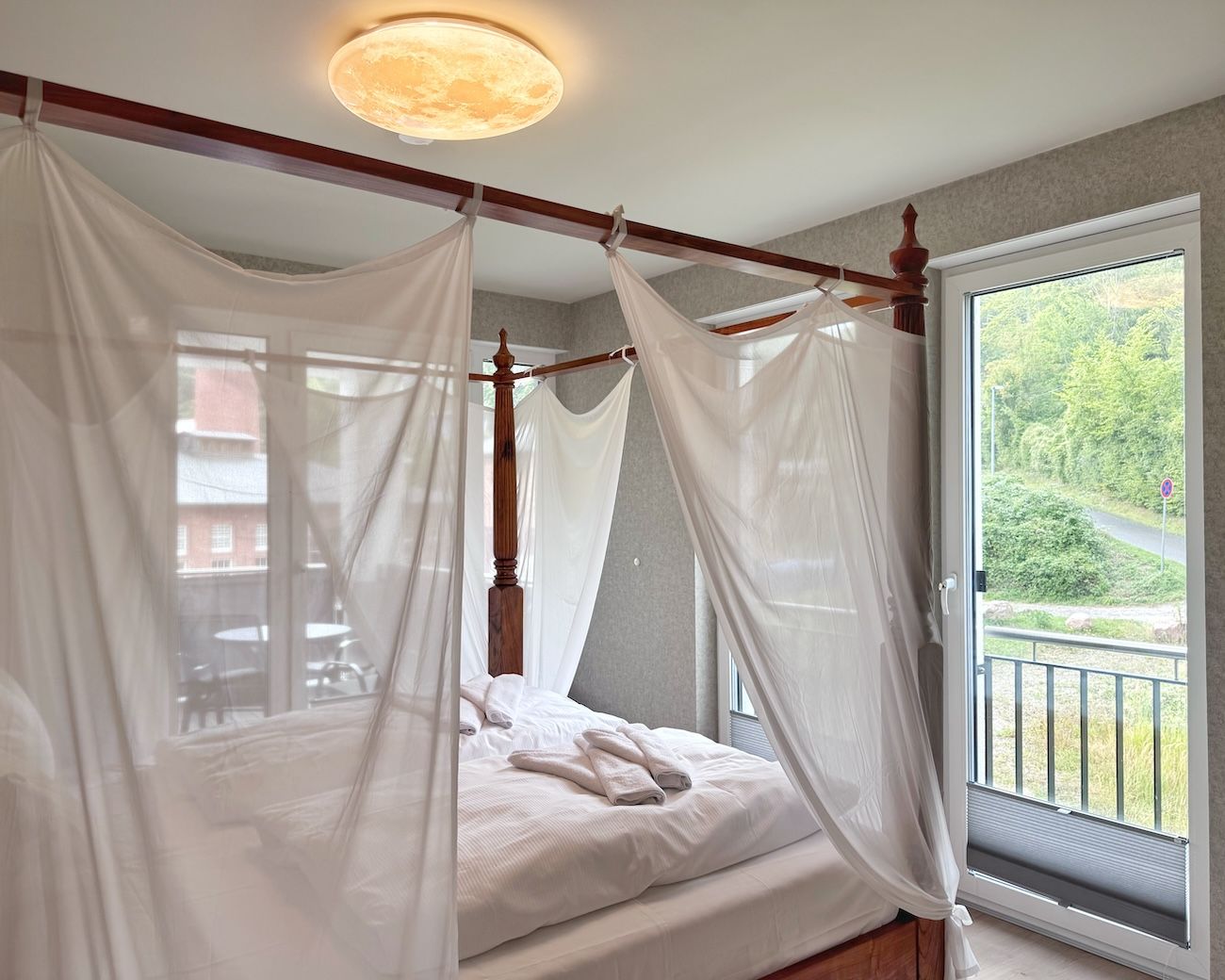A four-poster canopy bed with white linens, a wooden headboard, and a view of greenery through a balcony door.