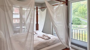 Uma cama com dossel e cortinas brancas, cabeceira de madeira, vista para o verde através de uma porta de varanda.