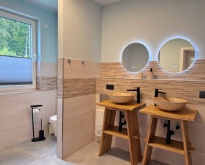 Ein modernes Badezimmer mit zwei Waschtischen aus Holz, einer Toilette und einem Fenster mit Jalousien.