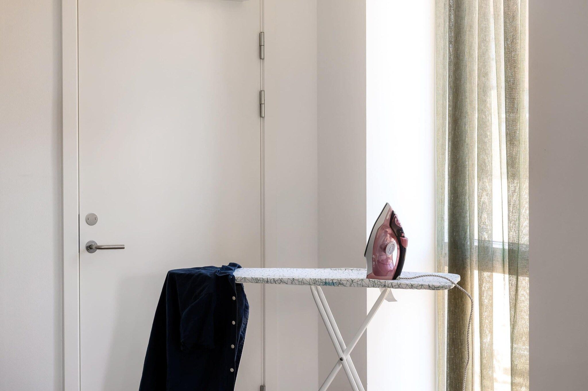 Una habitación con una puerta blanca, una tabla de planchar pequeña con un planchador, y una prenda azul oscuro colgada en la puerta.