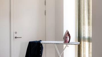 Una habitación con una puerta blanca, una tabla de planchar pequeña con un planchador, y una prenda azul oscuro colgada en la puerta.