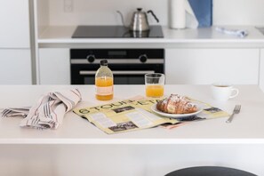 Ett frukostbord med en tidning, en flaska apelsinjuice, ett glas apelsinjuice, en croissant, en kopp kaffe och en gaffel.