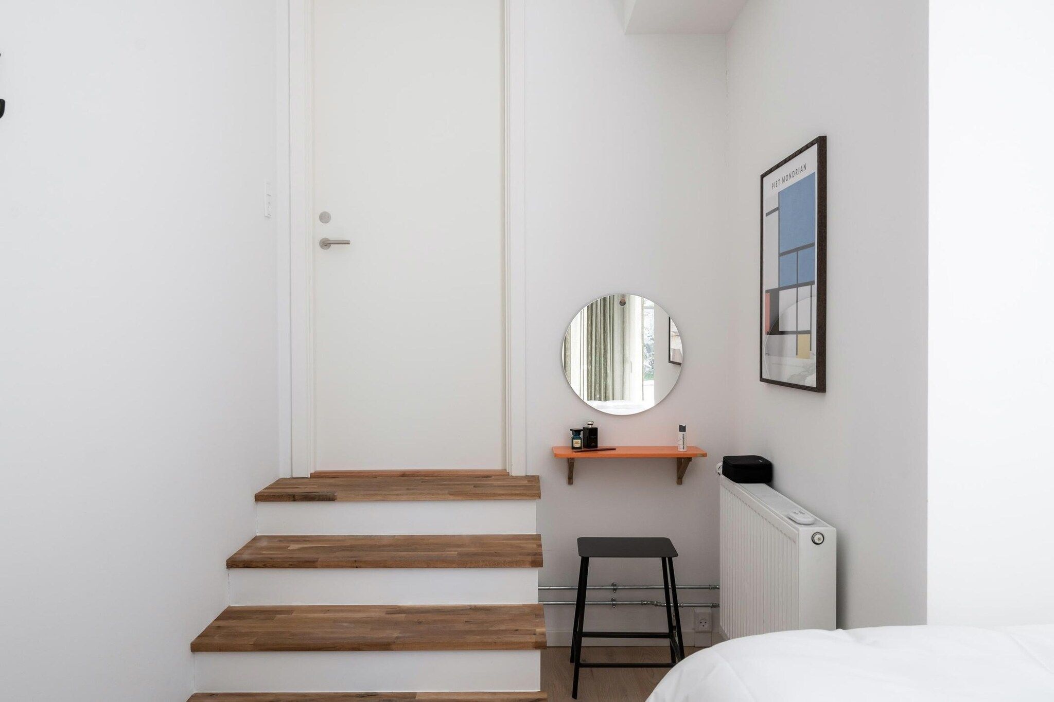 Un dormitorio con una puerta blanca, escalones de madera, un espejo redondo, un estante pequeño, un radiador y una cama.