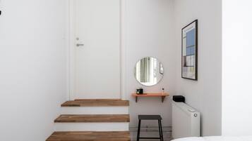 Un dormitorio con una puerta blanca, escalones de madera, un espejo redondo, un estante pequeño, un radiador y una cama.