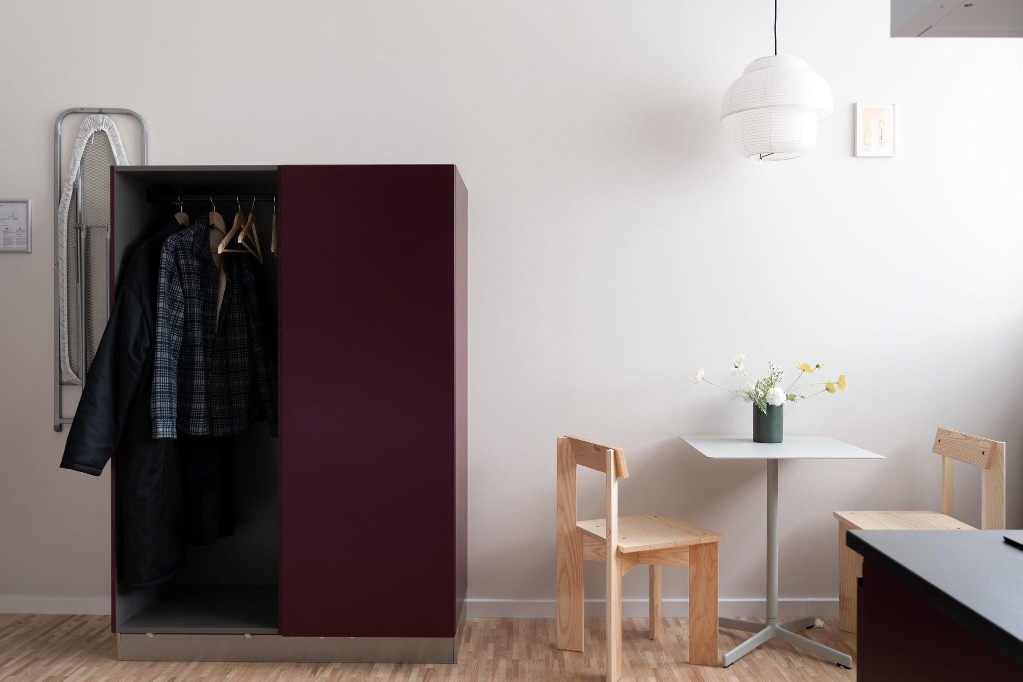 A room with a wardrobe, a table, two chairs, a hanging light fixture, and a small vase with flowers.