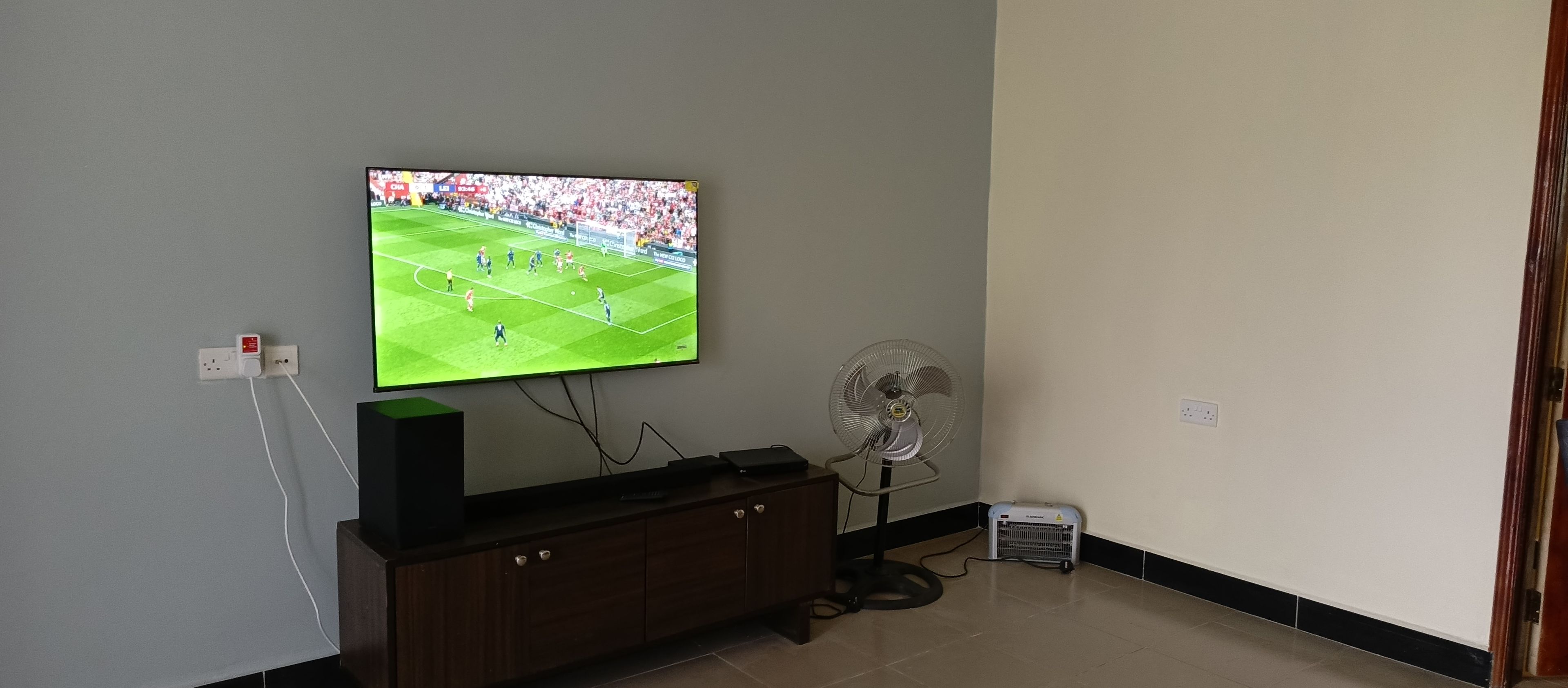 Ein Fernsehgerät an der Wand, das ein Sportspiel zeigt, ein Holzschrank darunter und ein Standventilator zur Rechten.
